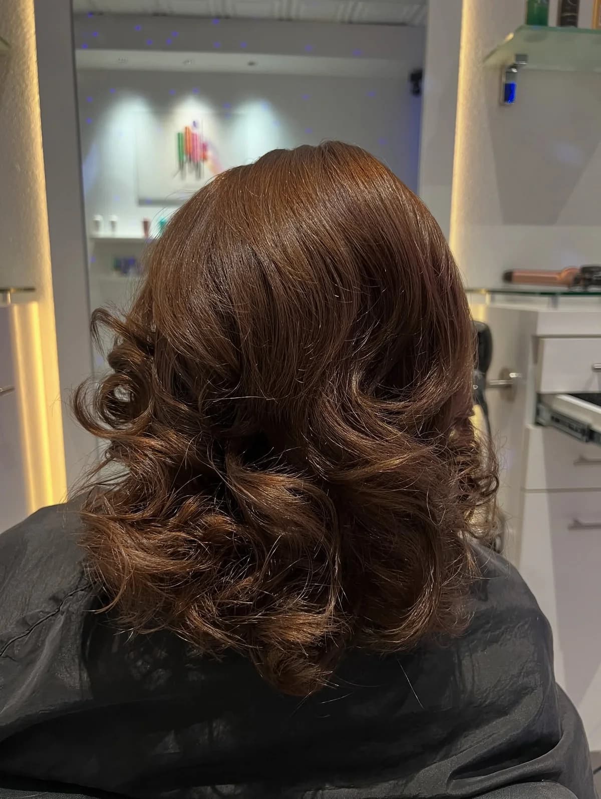 Warm chestnut brown hair styled in soft curls, photographed from behind in a salon